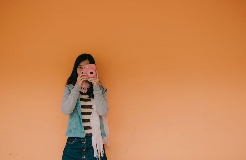 pexels photo 881633 Young woman holding a pink camera stands against an orange wall, exuding a playful fashion vibe.